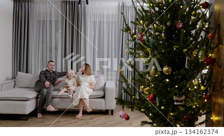 Cheerful family relaxing on couch beside sparkling christmas tree, sharing warmth and joy during seasonal celebration at home Cheerful family relaxing on couch beside sparkling christmas tree, sharing warmth and joy during seasonal celebration at home 134162374
