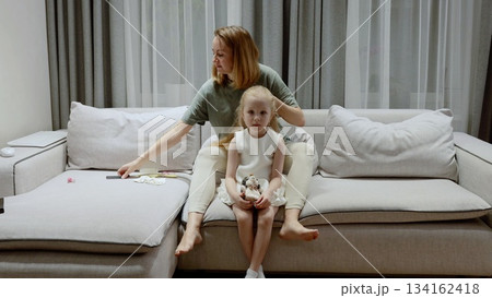 Mother gently brushing daughter's hair, sitting together on living room sofa, sharing intimate familial connection and tender care Mother gently brushing daughter's hair, sitting together on living room sofa, sharing intimate familial connection and tender care 134162418