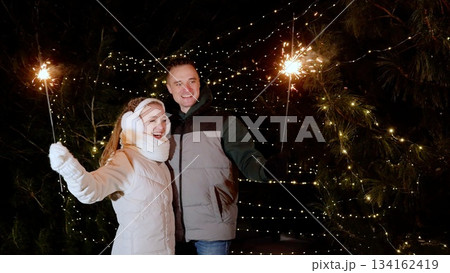 Celebrating winter holiday, couple standing outdoors at night, holding sparklers with glowing fir trees behind, sharing joyful moment during festive season Celebrating winter holiday, couple standing outdoors at night, holding sparklers with glowing fir trees behind, sharing joyful moment during festive season 134162419