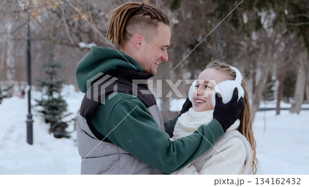 Couple sharing playful moment, dreadlocked man adjusting earmuffs for girlfriend during snowy winter park walk, both laughing 134162432
