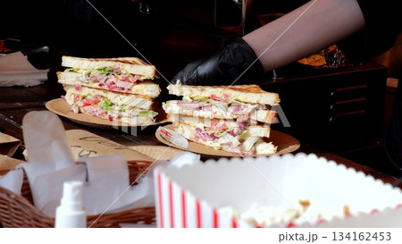 Vendor crafts multitiered deli sandwiches, Food stand displays stacked sandwiches with condiments and popcorn, Bustling street market scene with vendor preparing multilayer sandwiches and snack boxes 134162453