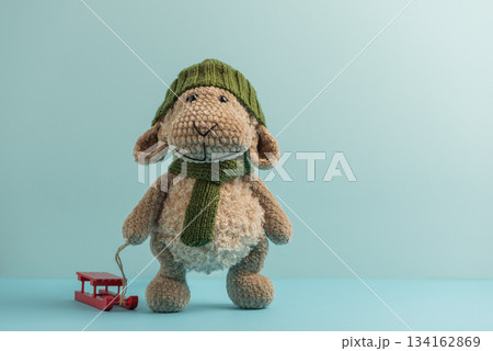 Plush Sheep in Green Hat and Scarf Pulling Red Sled on Blue Background 134162869