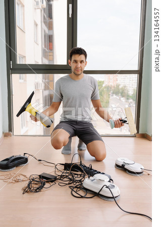 Vertical portrait of young man kneeling on floor, holding cordless window vacuum cleaner and wiper, preparing robot window cleaners and other appliances for cleaning windows, looking at camera. 134164557