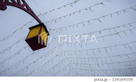 Decorative street lantern against a gray winter sky with a ceiling of suspended holiday light cables and wires 134164559