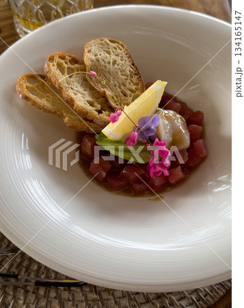 Beautifully decorated tuna tartare dish with avocado and scallop slices, lemon, bread chips and decorated with flowers on a white plate 134165147