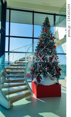 A modern interior with a large decorated Christmas tree next to a curved glass staircase in a bright room. The concept of Christmas in contemporary design, festivity, and a seasonal atmosphere. 134165204