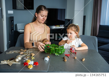 Mother and her little daughter decorating a Christmas wreath together at home. Strengthening family bonds through shared festive traditions and creative seasonal activities. Mother and her little daughter decorating a Christmas wreath together at home. Strengthening family bonds through shared festive traditions and creative seasonal activities. 134165751