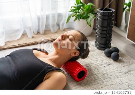 Young woman lying on the floor using a small red foam roller to massage her neck and occipital area. Daily self care routine to relieve muscle tension and improve cervical spine health. Young woman lying on the floor using a small red foam roller to massage her neck and occipital area. Daily self care routine to relieve muscle tension and improve cervical spine health. 134165753