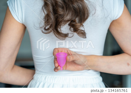 Back view of a woman holding a pink menstrual cup behind her back. Emphasizing sustainable period care, privacy, and eco-conscious personal hygiene choices. 134165761