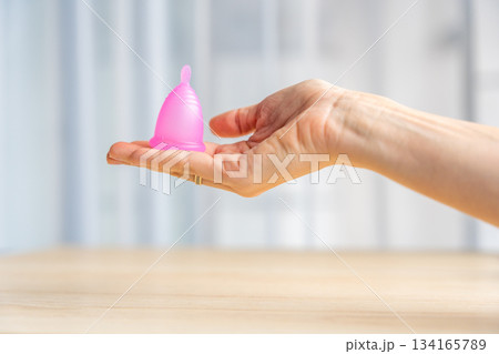 Close-up of a pink menstrual cup standing upright on a woman open palm over wooden table. Feminine hygiene product promoting zero waste, comfort, and sustainable period care. Close-up of a pink menstrual cup standing upright on a woman open palm over wooden table. Feminine hygiene product promoting zero waste, comfort, and sustainable period care. 134165789