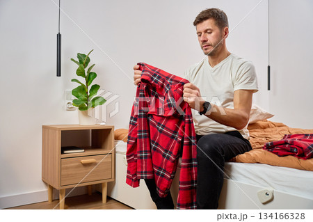 Man sitting on bed and holding plaid pajamas in bedroom at home 134166328