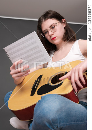 Woman musician holds sheet music in front of her, deciphering and analyzing new musical creation in creative process during rehearsal. Young guitarist singer songwriter with guitar wearing eyeglasses 134166374