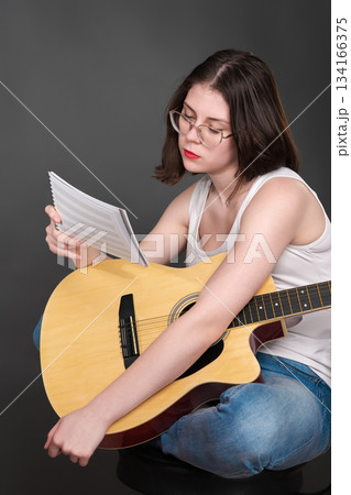 Musician is puzzled, holding musical notes in front as she figures out and breaks down new piece of music. Singer songwriter with guitar on lap. She in middle of creative process during rehearsal 134166375