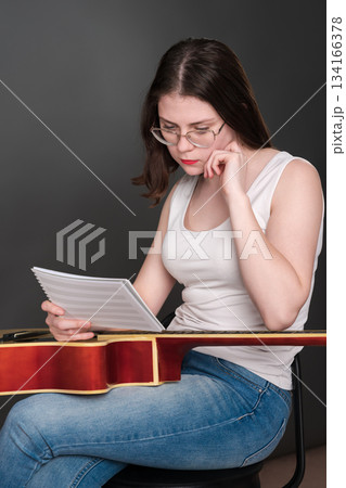 Female guitar player, intrigued by intricacies of her craft, holds musical notes before her, contemplating and interpreting novel musical composition. Singer songwriter in midst of creative process 134166378