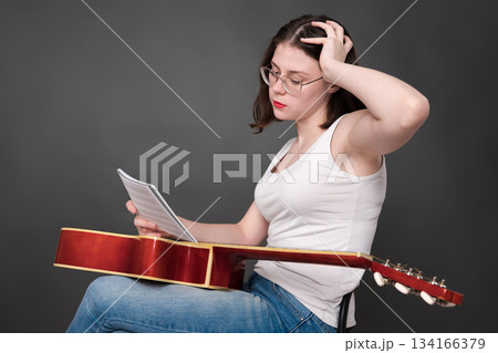 Perplexed female musician holds musical notes in front of her, deciphering and analyzing new musical creation. Female singer songwriter sitting with guitar on lap in creative process during rehearsal 134166379