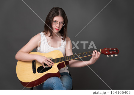 Woman guitarist playing classical six string acoustic guitar and looking at camera. 18 year old woman guitarist of Caucasian ethnicity, part of Generation Z, wearing eyeglasses, white tank top, jeans 134166403
