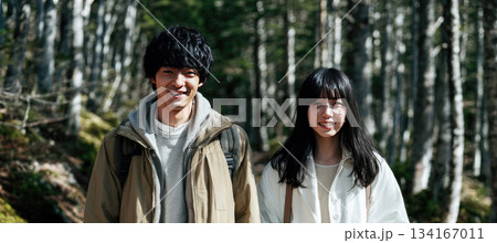 Young Japanese couple walking forest trail, smiling, wearing casual jackets, surrounded by tall trees and natural sunlight, peaceful outdoor atmosphere, spring or autumn season 134167011