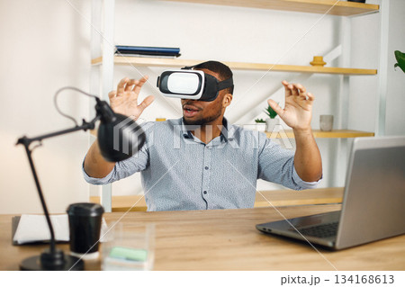 Black business man sitting in office with a laptop. Bearded man using virtual reality glases. Man wearing blue shirt. Black business man sitting in office with a laptop. Bearded man using virtual reality glases. Man wearing blue shirt. 134168613
