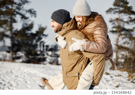 Portrait of romantic couple having fun on winter walk. Man carrying woman piggyback. Family wearing warm jacket and knitted hats. Portrait of romantic couple having fun on winter walk. Man carrying woman piggyback. Family wearing warm jacket and knitted hats. 134168642