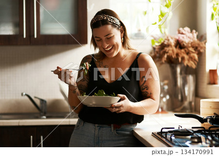 Woman, diet and person eating salad in her home kitchen and is happy for a meal with nutrition or healthy lunch. Smile, food and young female vegan in her apartment or house and eat vegetables 134170991