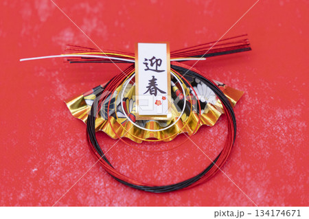 「迎春」の札が付いた扇型の水引飾りと布地のような赤い背景 134174671