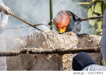 Blacksmith holding a hot crucible by the iron pliers from furnace and pouring the melting gold into the statue block. Blacksmith holding a hot crucible by the iron pliers from furnace and pouring the melting gold into the statue block. 134174952