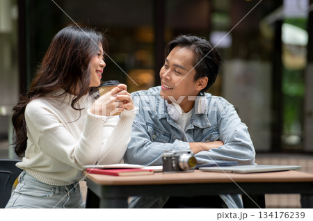 Young asian couple a man looking at woman holding coffee sitting at outdoor table in front of a cafe Young asian couple a man looking at woman holding coffee sitting at outdoor table in front of a cafe 134176239