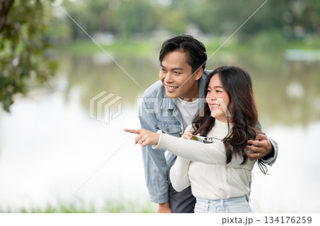 Young asian couple a woman pointing aside man hugging her shoulder while sitting outdoors in park Young asian couple a woman pointing aside man hugging her shoulder while sitting outdoors in park 134176259