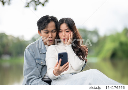 Young asian couple a man hugging woman taking selfie picture on phone while sitting outdoors in park Young asian couple a man hugging woman taking selfie picture on phone while sitting outdoors in park 134176266