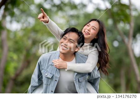 Young asian couple woman pointing on man's shoulder while getting piggyback walking outdoors in park 134176270