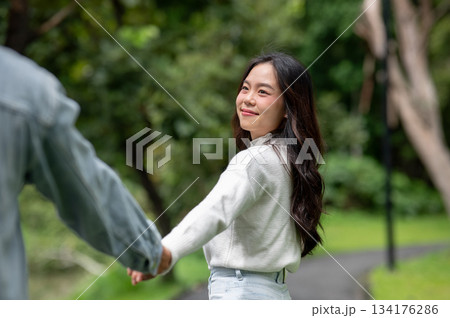 Young couple holding hands woman turning looking back at man while walking outdoors on road in park 134176286