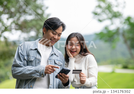 Young asian couple man looking at phone and woman holding credit card while standing outdoor in park 134176302