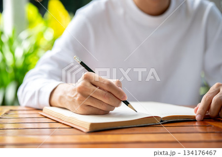 Close up of a man's hand holding pencil writing in notebook while sitting outdoors at wooden table. Close up of a man's hand holding pencil writing in notebook while sitting outdoors at wooden table. 134176467