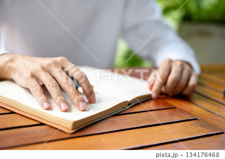 Close up of a man's hand holding pencil over notebook on wooden table while sitting outdoors in cafe Close up of a man's hand holding pencil over notebook on wooden table while sitting outdoors in cafe 134176468