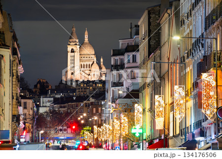Illuminated Sacre-Coeur Basilica rises above city street at night in Paris, France. View from Saint-Ouen features festive Christmas lights and apartment buildings 134176506