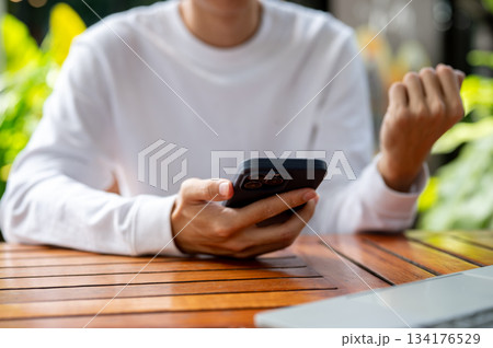 Close up of man's hand holding phone making a fist aside laptop sitting outdoor at wooden cafe table Close up of man's hand holding phone making a fist aside laptop sitting outdoor at wooden cafe table 134176529