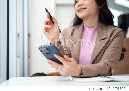Asian business woman office worker holding pen and phone thinking over notebook sits at cafe table. 134176835