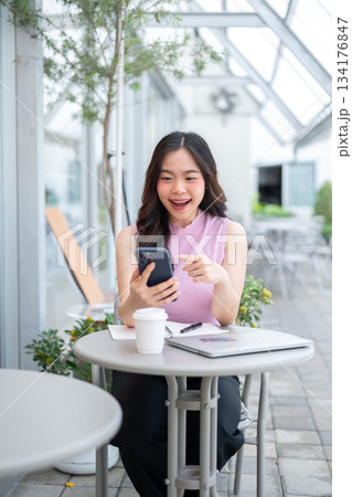 Asian woman student holding and laughing at phone while sitting with laptop and coffee at cafe table Asian woman student holding and laughing at phone while sitting with laptop and coffee at cafe table 134176847