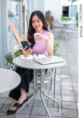 Asian woman student holding coffee and looking at phone sitting with laptop and coffee at cafe table Asian woman student holding coffee and looking at phone sitting with laptop and coffee at cafe table 134176848