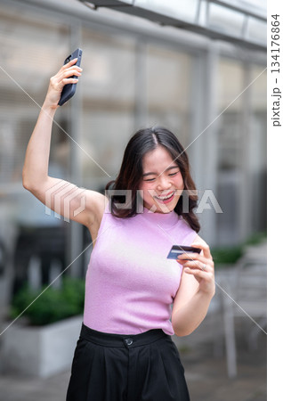 Young asian woman looking at credit card raising a hand with phone while standing in front of a cafe Young asian woman looking at credit card raising a hand with phone while standing in front of a cafe 134176864