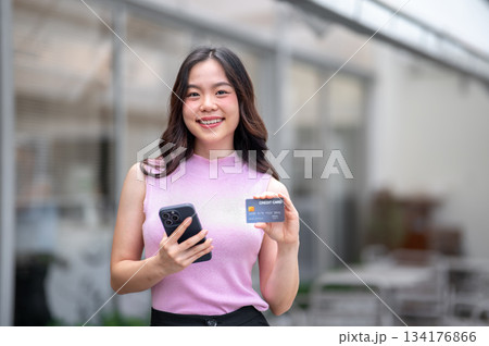 Young asian woman holding phone and showing a credit card in hand while standing in front of a cafe 134176866