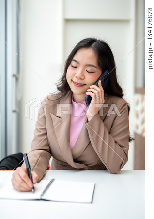 Asian businesswoman or office worker writing in notebook and talking on phone sitting at cafe table. 134176878