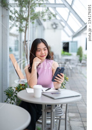 Asian woman student or freelancer holding and looking at phone aside coffee and laptop on cafe table Asian woman student or freelancer holding and looking at phone aside coffee and laptop on cafe table 134176891