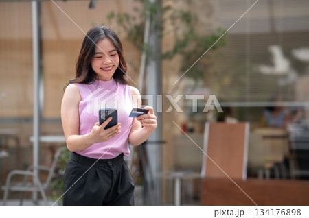 Asian young woman holding credit card looking at phone with smile while standing in front of a cafe. 134176898