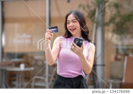 Smiling asian young woman holding phone looking at credit card while standing in front of the cafe. Smiling asian young woman holding phone looking at credit card while standing in front of the cafe. 134176899