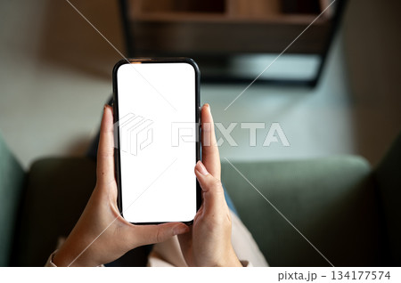 Close up a woman hands holding white screen smartphone while sitting on green sofa in cafe or lounge Close up a woman hands holding white screen smartphone while sitting on green sofa in cafe or lounge 134177574