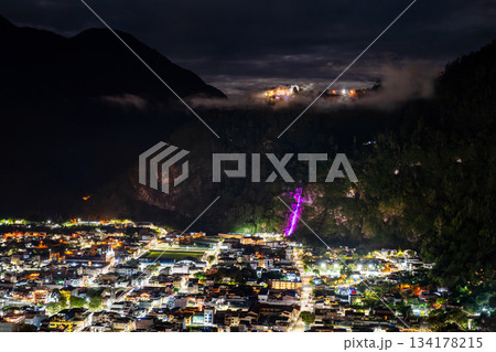 Banos de Agua Santa town center glows at night in Ecuador. Panoramic view features illuminated Cascada de la Virgen waterfall on cliffside above city streets Banos de Agua Santa town center glows at night in Ecuador. Panoramic view features illuminated Cascada de la Virgen waterfall on cliffside above city streets 134178215