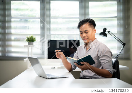 Asian businessman or office worker holding pen and looking at notebook aside laptop on working table 134178220