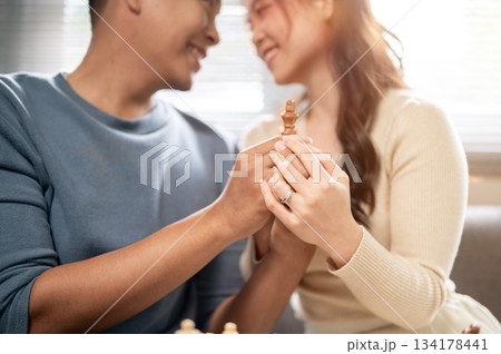 Close up smiling asian man and woman holding a wooden chess piece together as sitting on home's sofa 134178441