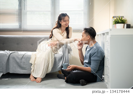 Asian man holding and kissing a woman's hand while holding coffee or tea cup and sitting aside sofa. Asian man holding and kissing a woman's hand while holding coffee or tea cup and sitting aside sofa. 134178444